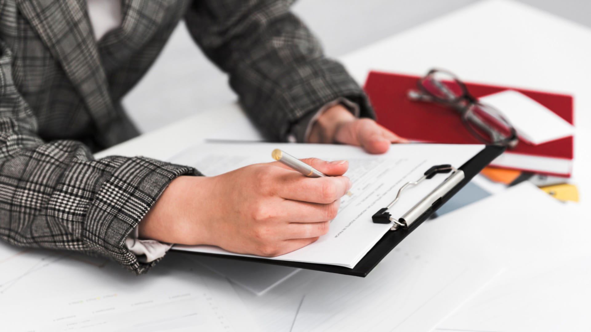 A person writes on a clipboard at a desk, surrounded by documents, files and a pair of glasses.